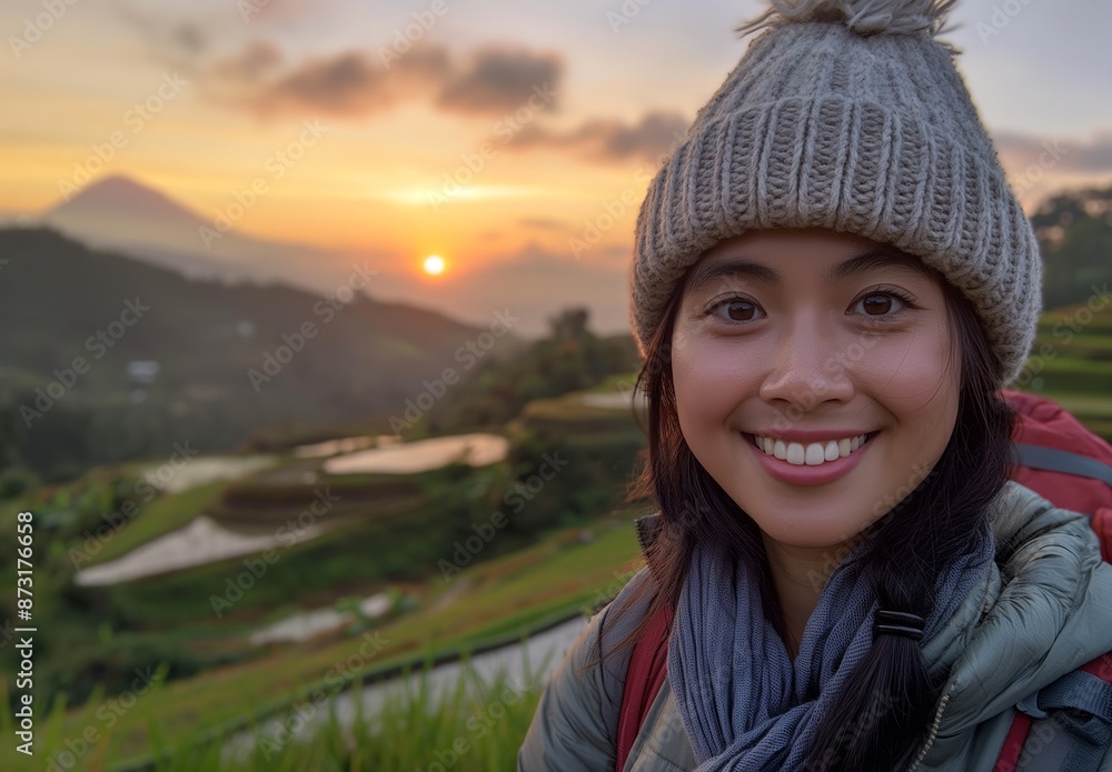 Portrait of an Asian woman wearing winter hat and jacket, smiling at the camera with rice terrace fields in background during sunset. Young asian girl traveler backpacker traveling adventure concept. Portrait of an Asian woman wearing winter hat and jacket, smiling at the camera with rice terrace fields in background during sunset. Young asian girl traveler backpacker traveling adventure concept.