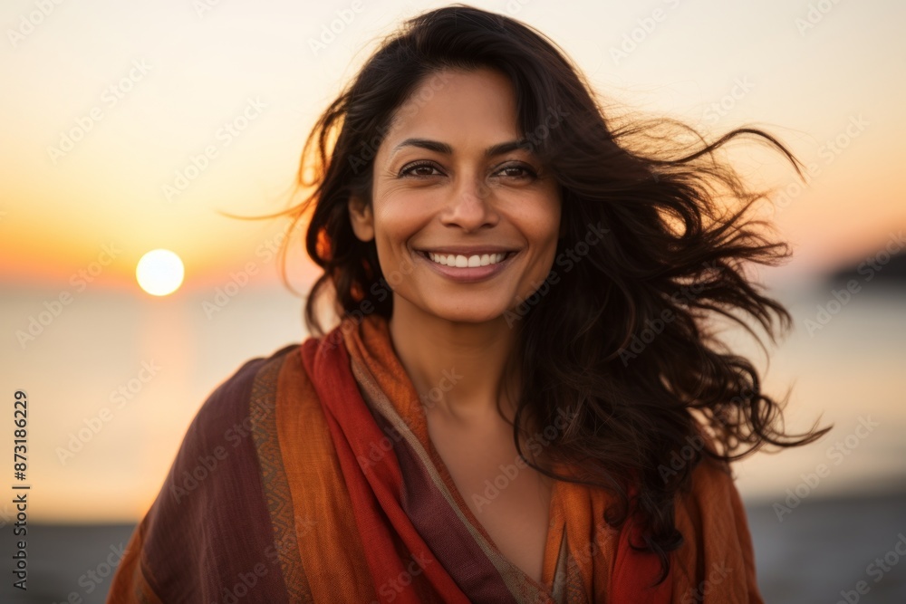 Fototapeta premium Portrait of a grinning indian woman in her 30s wearing a versatile buff over vibrant beach sunset background