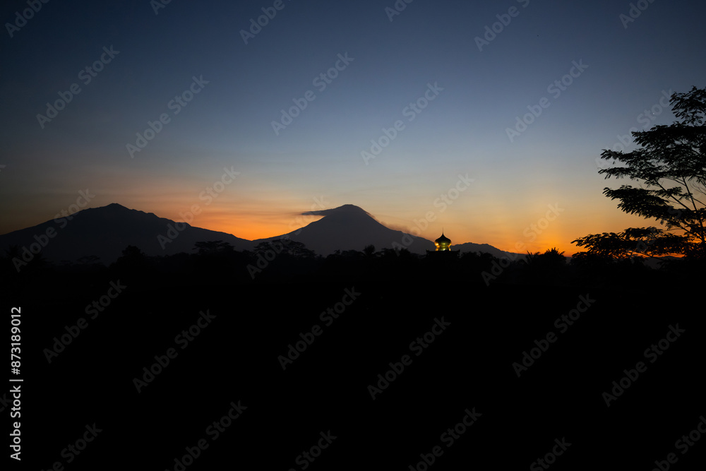 Sunrise view from Borobudur, Magelang, Indonesia towards Mount Merapi and Mount Merbabu. Mosque spire minoret visible, clouds brush mountain top, trees in silhouette, golden light on horizon. Sunrise view from Borobudur, Magelang, Indonesia towards Mount Merapi and Mount Merbabu. Mosque spire minoret visible, clouds brush mountain top, trees in silhouette, golden light on horizon.