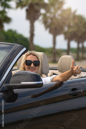 Portrait of a young smiling blonde woman in sunglasses driving a convertible convertible showing a thumbs up. Concept of travel, vacation, rental. Vertical photo