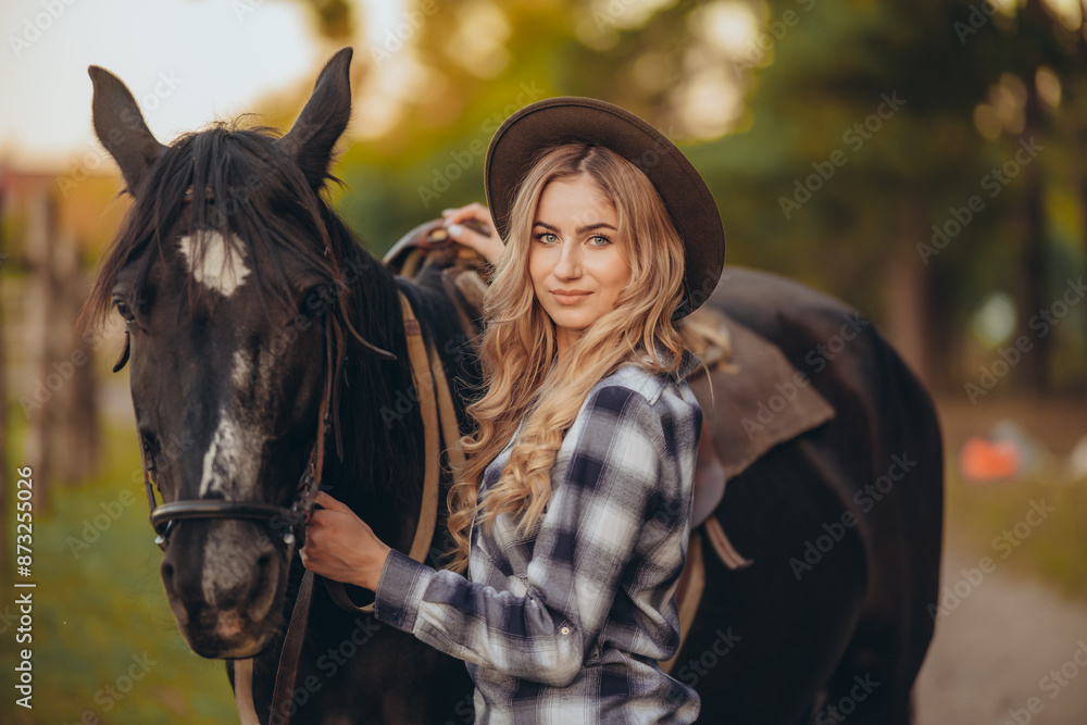Obraz premium A young woman with a horse stands near a fence in autumn at sunset. Breeding thoroughbred horses for equestrian sport.