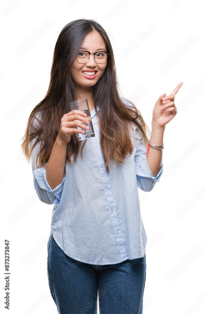 Young asian woman drinking a glass of water over isolated background very happy pointing with hand and finger to the side Young asian woman drinking a glass of water over isolated background very happy pointing with hand and finger to the side