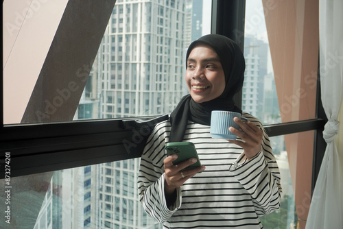 A woman is smiling and holding a cup of coffee while holding a cell phone. Concept of relaxation and enjoyment, as the woman is taking a break from her daily routine to enjoy a warm beverage