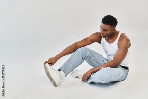 Fotografie A young and stylish African American man sits calmly on the ground in casual attire of jeans and a tank top