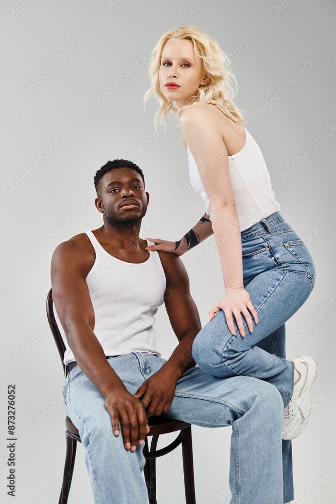 A young interracial couple sitting comfortably on chairs, engrossed in conversation, against a neutral grey backdrop. A young interracial couple sitting comfortably on chairs, engrossed in conversation, against a neutral grey backdrop.