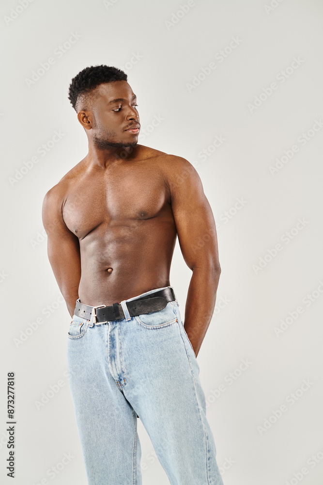 Fototapeta premium A young, shirtless, African American man confidently posing in a studio against a grey background.