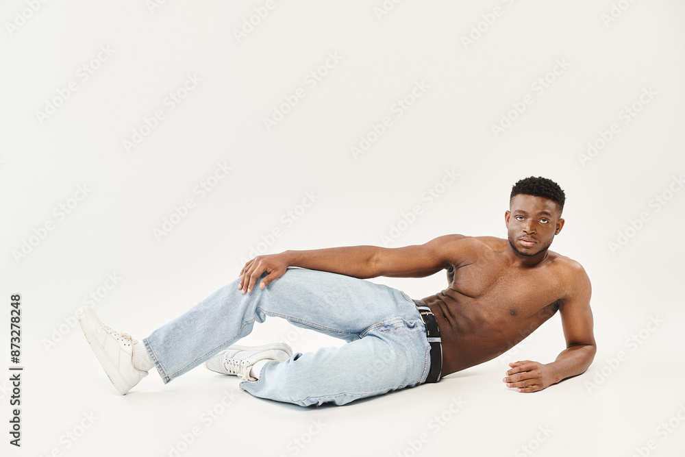 Fototapeta premium A young shirtless African American man strikes a captivating pose on the ground in a studio setting against a grey background.