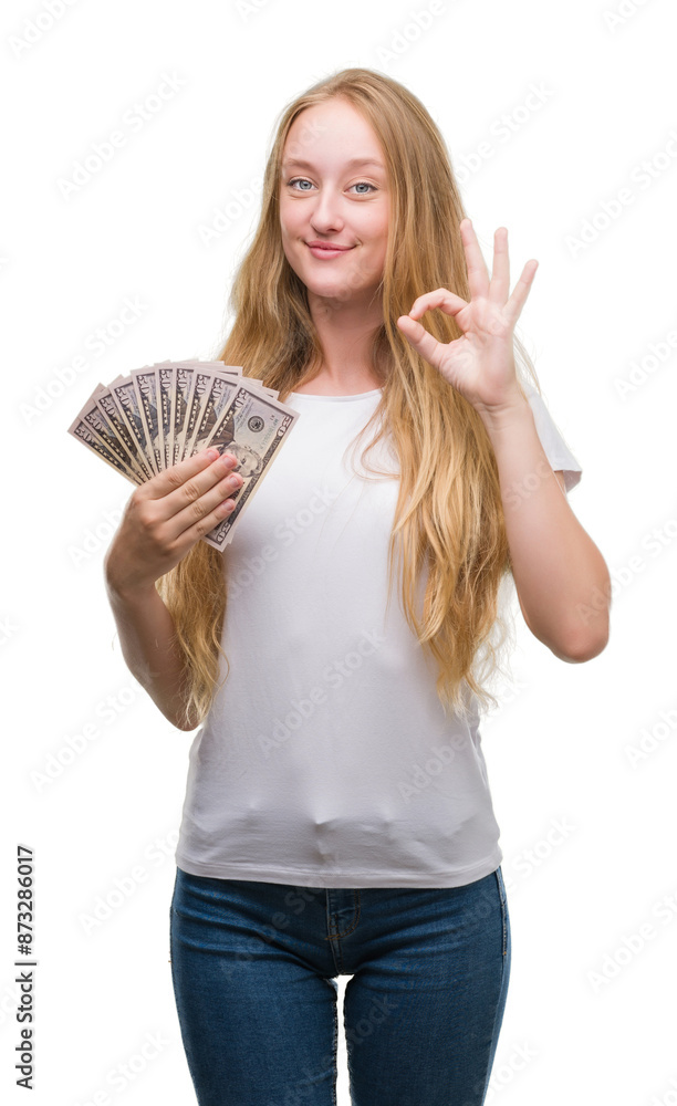 Blonde teenager woman holding dollars doing ok sign with fingers, excellent symbol Blonde teenager woman holding dollars doing ok sign with fingers, excellent symbol