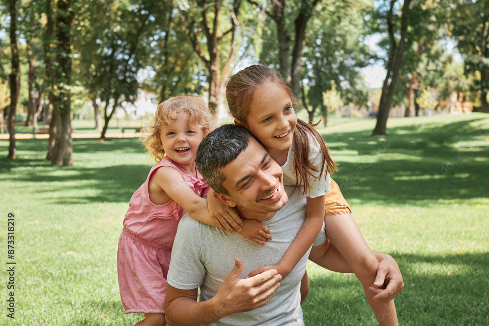 Father and daughters love. Kid playing in nature. Happy family outdoors. Cheerful summer park fun. Laughing father carrying his happy joyful daughters on back outdoors Father and daughters love. Kid playing in nature. Happy family outdoors. Cheerful summer park fun. Laughing father carrying his happy joyful daughters on back outdoors