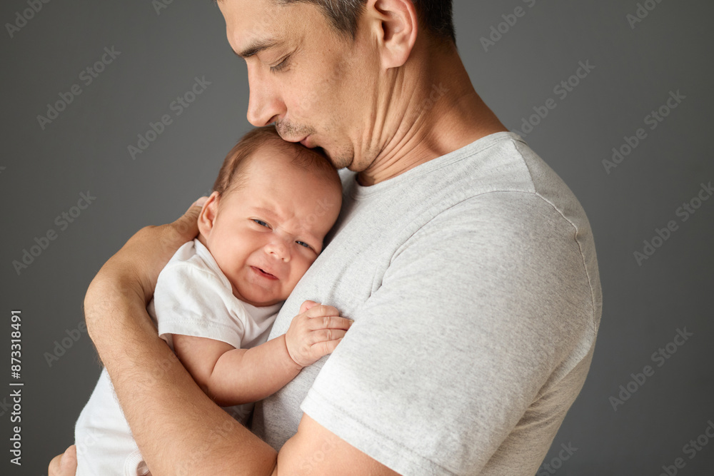 Loving Caucasian father holding his newborn baby in his arms isolated over gray background young man wearing white T-shirt kissing his infant son with love and gentle Loving Caucasian father holding his newborn baby in his arms isolated over gray background young man wearing white T-shirt kissing his infant son with love and gentle