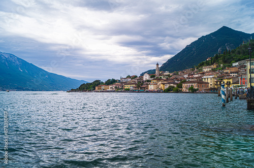 A view of Limone on Lake Garda. The lake front buildings can be seen, with the mountain rising above it. The sky is overcast but clouds are beginning to break up, brightening the scene.