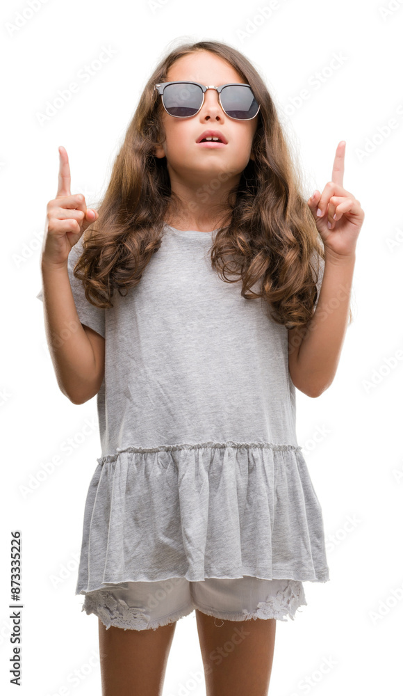 Brunette hispanic girl wearing sunglasses amazed and surprised looking up and pointing with fingers and raised arms. Brunette hispanic girl wearing sunglasses amazed and surprised looking up and pointing with fingers and raised arms.