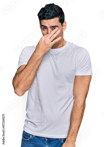 Handsome hispanic man wearing casual white t shirt smelling something stinky and disgusting, intolerable smell, holding breath with fingers on nose. bad smell