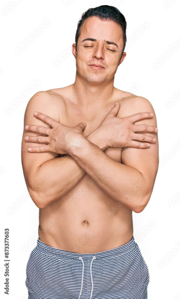 Fototapeta premium Handsome young man wearing swimwear shirtless hugging oneself happy and positive, smiling confident. self love and self care
