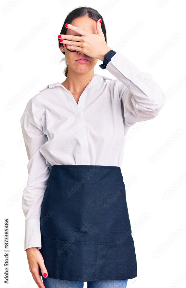Beautiful brunette young woman wearing professional waitress apron covering eyes with hand, looking serious and sad. sightless, hiding and rejection concept Beautiful brunette young woman wearing professional waitress apron covering eyes with hand, looking serious and sad. sightless, hiding and rejection concept