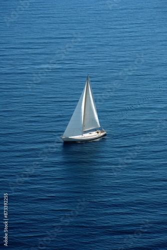 A lone sailboat with white sails glides smoothly across the calm, deep blue sea, creating a serene and tranquil scene