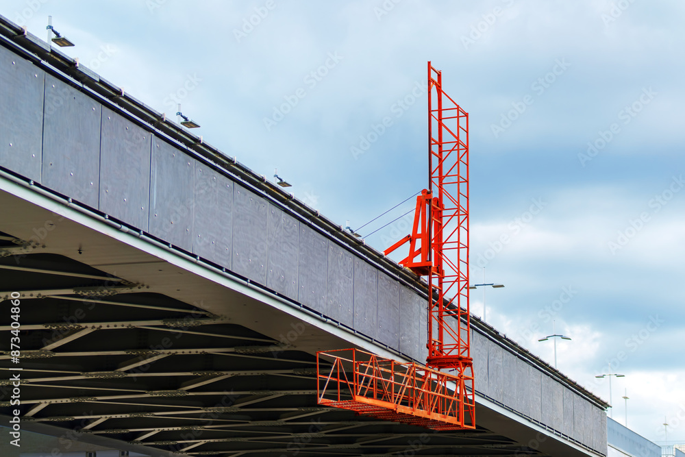 Orange bridge maintenance platform on a granite bridge on a cloudy day. Signifies infrastructure upkeep, engineering, urban maintenance Orange bridge maintenance platform on a granite bridge on a cloudy day. Signifies infrastructure upkeep, engineering, urban maintenance