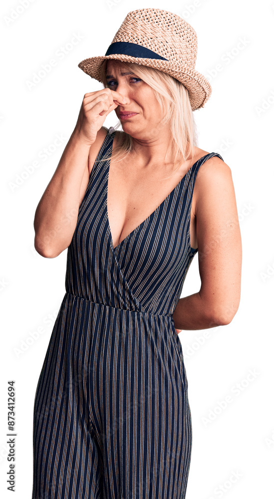 Young beautiful blonde woman wearing summer hat and dress smelling something stinky and disgusting, intolerable smell, holding breath with fingers on nose. bad smell Young beautiful blonde woman wearing summer hat and dress smelling something stinky and disgusting, intolerable smell, holding breath with fingers on nose. bad smell