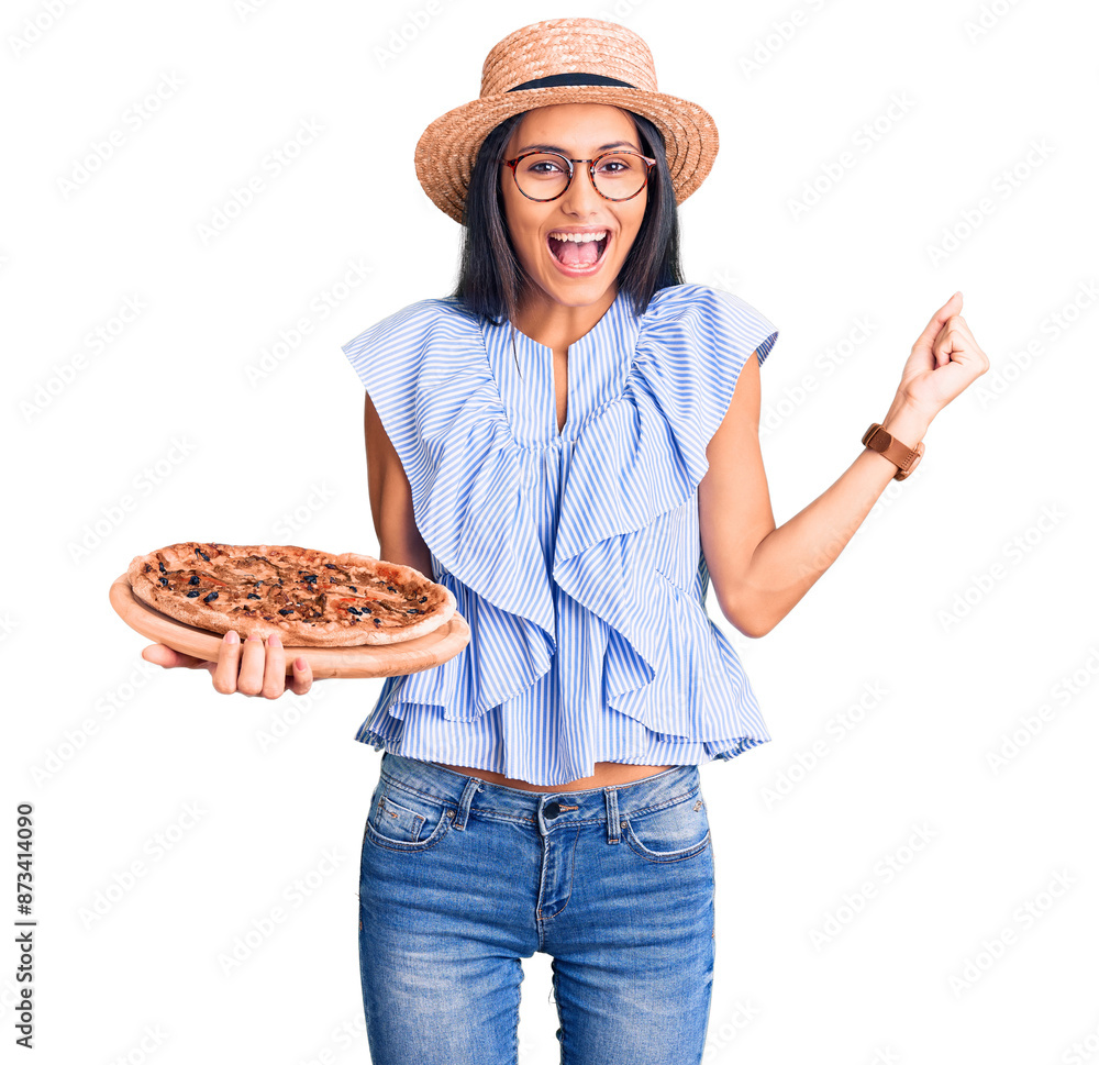 Young beautiful latin girl wearing summer hat and glasses holding pizza screaming proud, celebrating victory and success very excited with raised arms Young beautiful latin girl wearing summer hat and glasses holding pizza screaming proud, celebrating victory and success very excited with raised arms