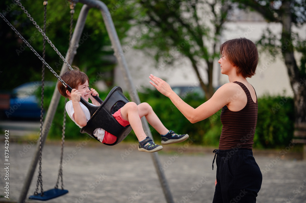Obraz premium A mother pushing her laughing young son on a swing at the playground, capturing a moment of pure joy and playful bonding together outdoors.