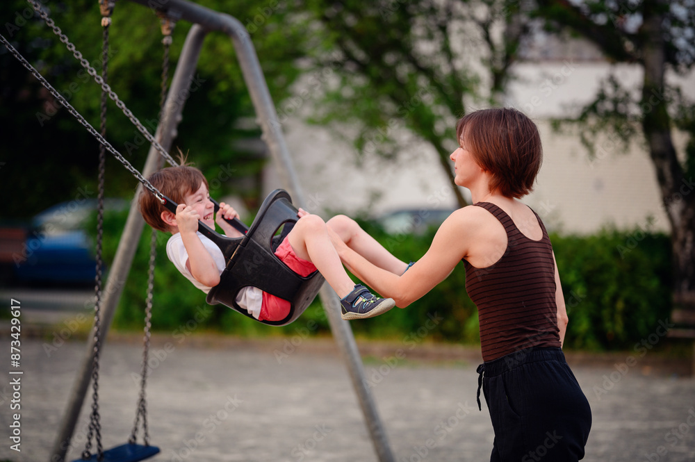 Obraz premium A mother pushing her laughing young son on a swing at the playground, capturing a moment of pure joy and playful bonding together outdoors.