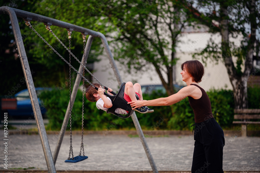 Obraz premium A mother pushing her son on a swing at the playground, both smiling and enjoying a joyful moment of play and bonding outdoors.