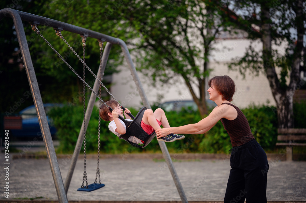 Obraz premium A mother pushing her son on a swing at the playground, both smiling and enjoying a joyful moment of play and bonding outdoors.