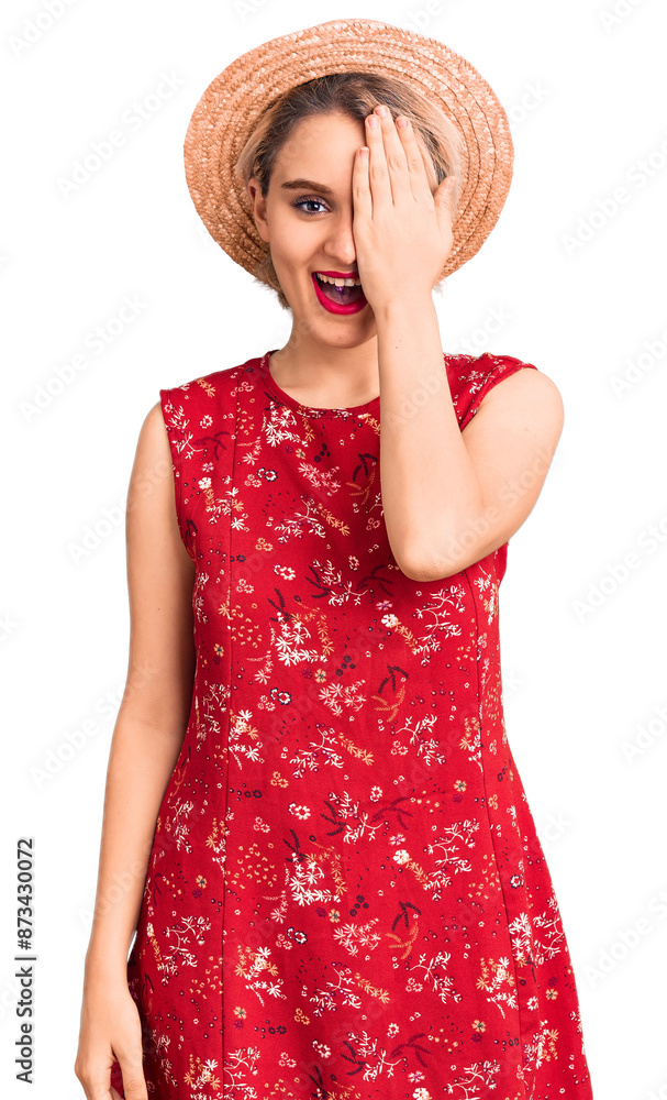 Young beautiful blonde woman wearing summer hat covering one eye with hand, confident smile on face and surprise emotion. Young beautiful blonde woman wearing summer hat covering one eye with hand, confident smile on face and surprise emotion.