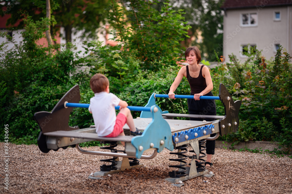 A mother and her young son enjoy playing on a seesaw at the playground. The boy sits on one end, looking ahead, while the mother balances the other side, both smiling and enjoying the moment. A mother and her young son enjoy playing on a seesaw at the playground. The boy sits on one end, looking ahead, while the mother balances the other side, both smiling and enjoying the moment.
