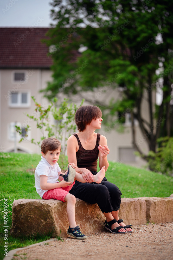 A mother and her young son sit together on a large rock in a park. The boy examines his shoe closely while the mother watches, capturing a moment of shared curiosity and care in the outdoors. A mother and her young son sit together on a large rock in a park. The boy examines his shoe closely while the mother watches, capturing a moment of shared curiosity and care in the outdoors.