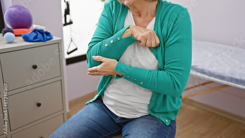 Mature woman holding elbow in pain at a physical therapy clinic, showing signs of discomfort and injury
