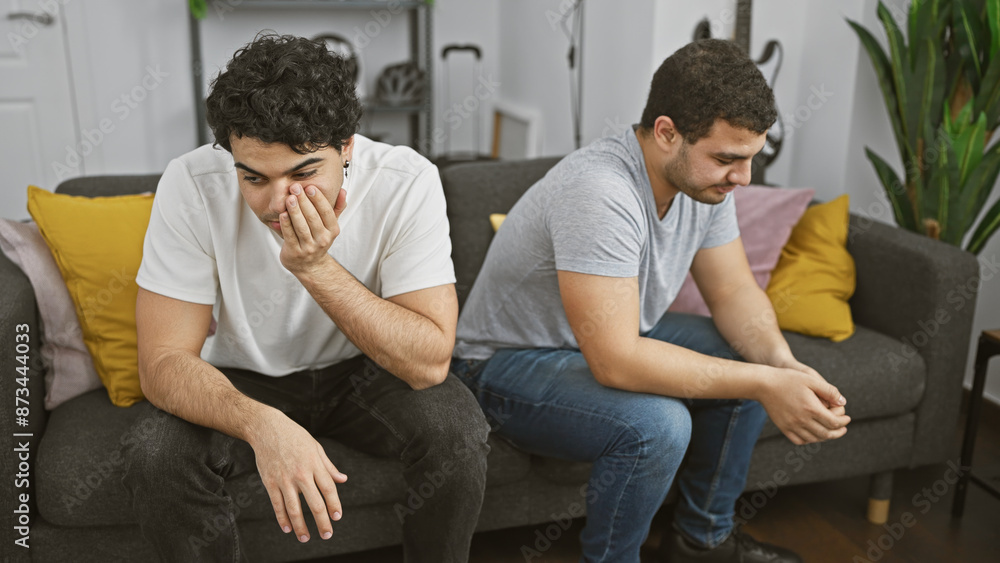 Two hispanic men showing distress while sitting apart on a couch indoors, conveying a tense atmosphere. Two hispanic men showing distress while sitting apart on a couch indoors, conveying a tense atmosphere.