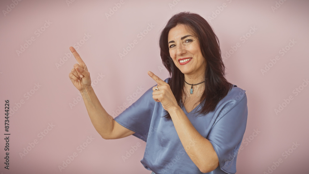 Smiling mature hispanic woman pointing upwards with both hands, against a soft pink background. Smiling mature hispanic woman pointing upwards with both hands, against a soft pink background.