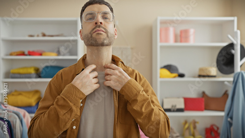 A stylish hispanic man adjusts his brown jacket in a bright modern wardrobe filled with assorted clothes.