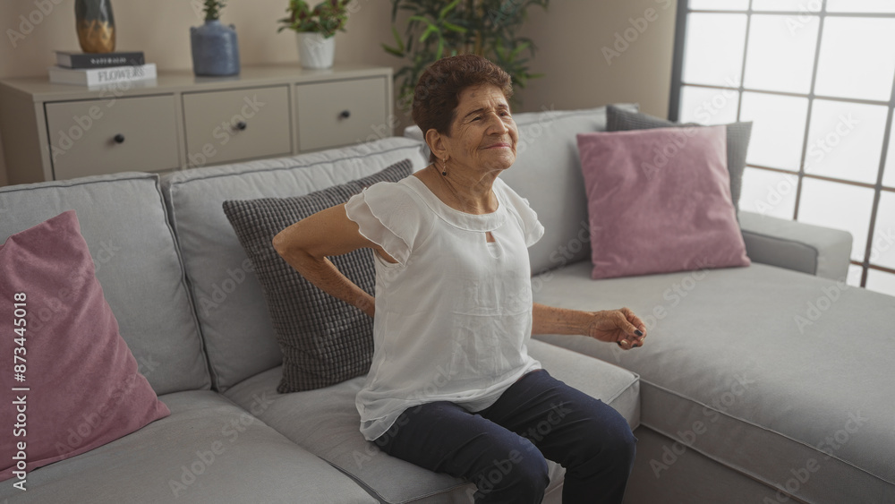 An elderly hispanic woman with short hair sits on a couch in a living room, experiencing back pain in a home environment. An elderly hispanic woman with short hair sits on a couch in a living room, experiencing back pain in a home environment.