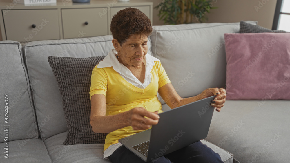 Elderly hispanic woman with short hair using a laptop at home in the living room on a comfortable sofa, engaged and focused on the device. Elderly hispanic woman with short hair using a laptop at home in the living room on a comfortable sofa, engaged and focused on the device.