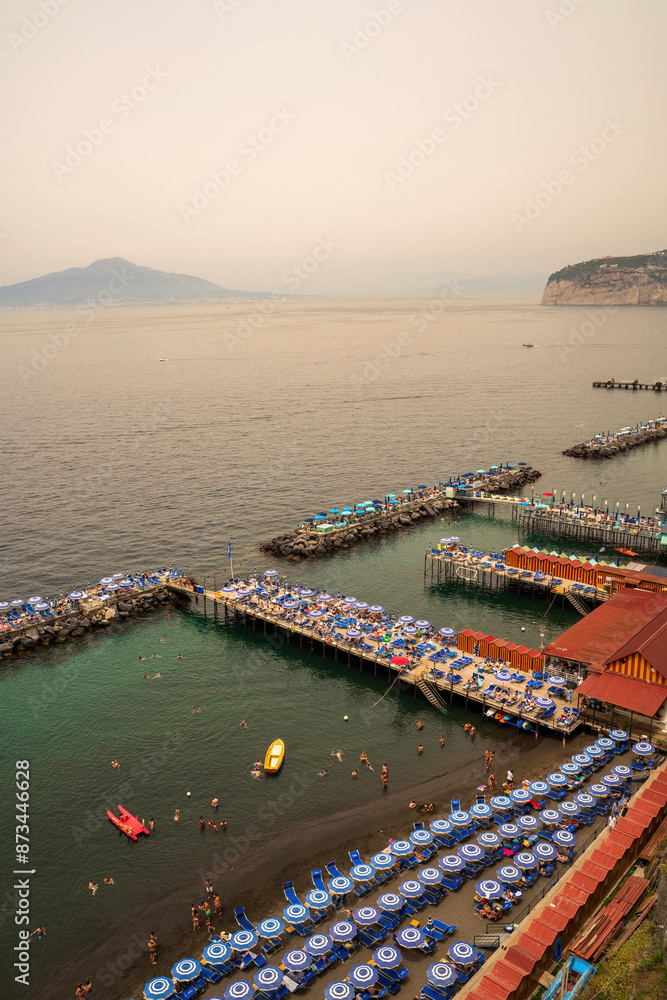 Sorrento, Italy - July 24, 2024: People relaxing on the beach on the pier and swimming in the sea on inflatable air beds in Sorrento and boats floating against mount vesuvius volcano