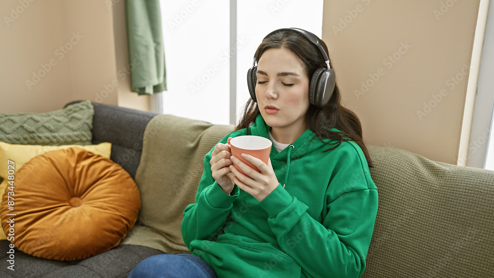 A young caucasian woman relaxes at home, enjoying music on headphones with eyes closed, savoring a cup of coffee in a cozy living room. A young caucasian woman relaxes at home, enjoying music on headphones with eyes closed, savoring a cup of coffee in a cozy living room.