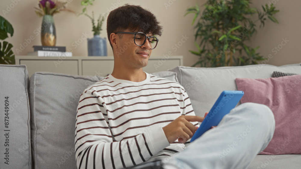Fototapeta premium Handsome young hispanic man relaxing indoors using a tablet in a modern living room, surrounded by cozy decor and houseplants.