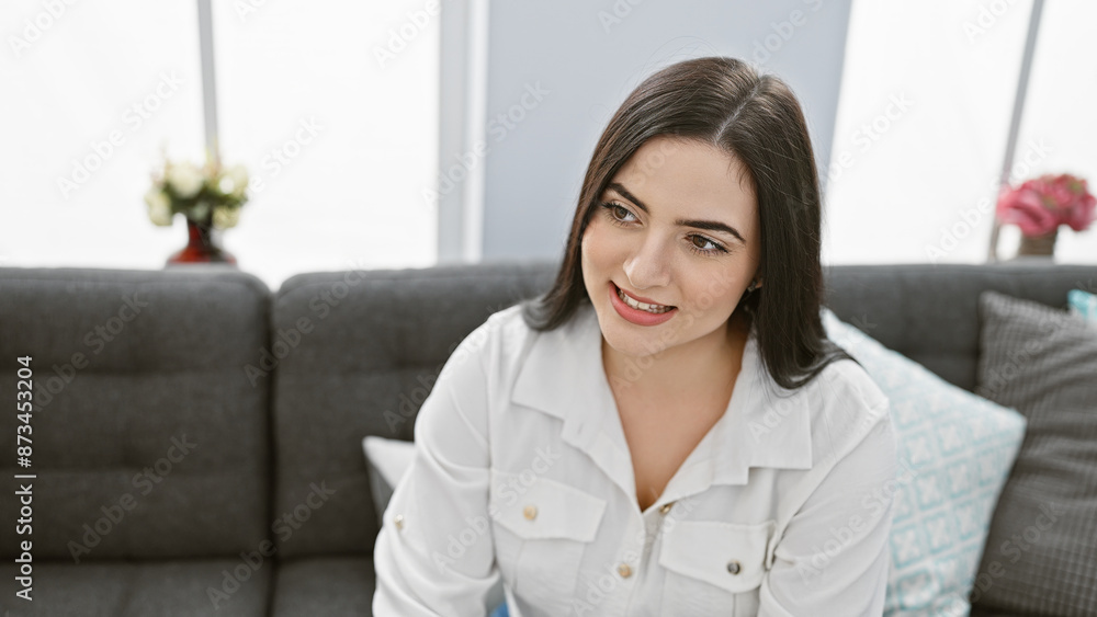 A smiling young hispanic woman seated comfortably in a stylish living room, exuding casual elegance and warmth. A smiling young hispanic woman seated comfortably in a stylish living room, exuding casual elegance and warmth.
