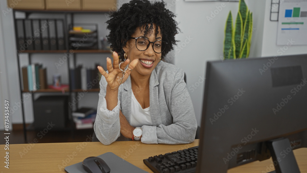 Obraz premium A beautiful young african american woman with curly hair, wearing glasses, smiles while sitting in an office holding keys, with a computer, indoor plant, and shelves in the background.