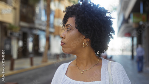 A beautiful young african american woman with curly hair standing on an urban city street, looking thoughtfully into the distance on a sunny day.