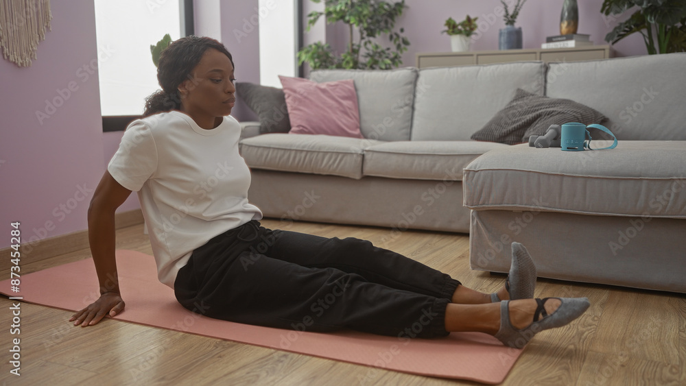 Young african american woman with curly hair doing stretching exercises on a yoga mat in a cozy living room with modern decor and wooden floor. Young african american woman with curly hair doing stretching exercises on a yoga mat in a cozy living room with modern decor and wooden floor.