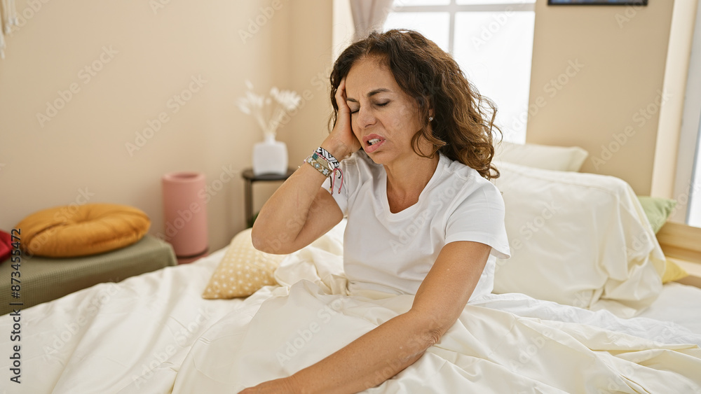 A middle-aged hispanic woman with curly hair looks stressed sitting on a bed in a cozy bedroom setting.