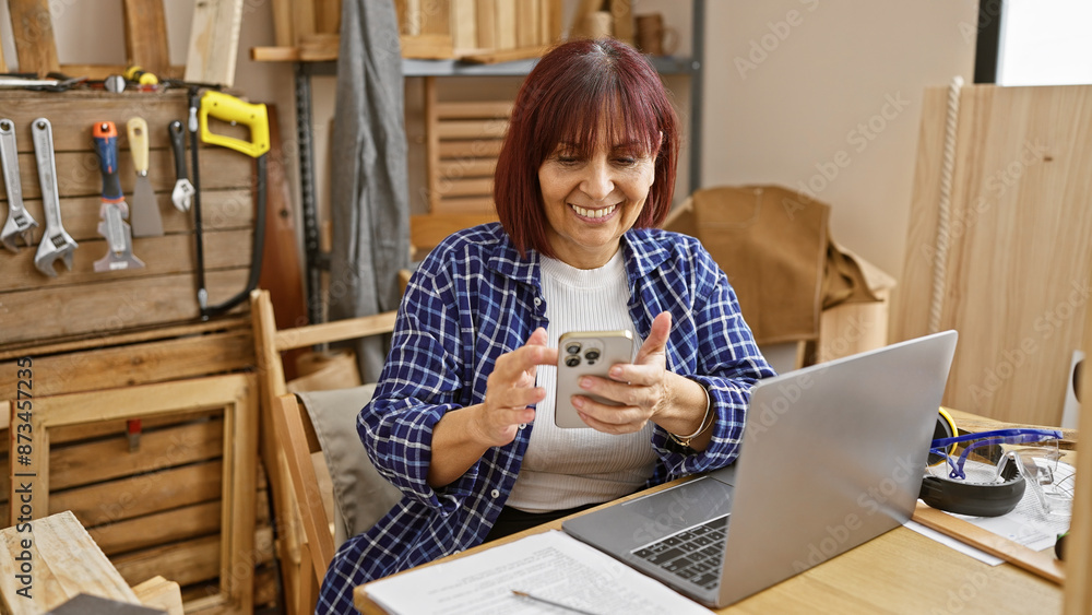 Smiling hispanic senior woman using smartphone in a carpentry workshop with laptop and tools Smiling hispanic senior woman using smartphone in a carpentry workshop with laptop and tools