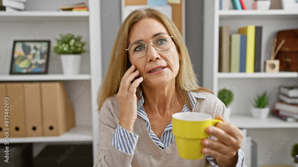 A pensive senior woman holding a coffee cup in a modern office, talking on the phone and gazing away thoughtfully. A pensive senior woman holding a coffee cup in a modern office, talking on the phone and gazing away thoughtfully.