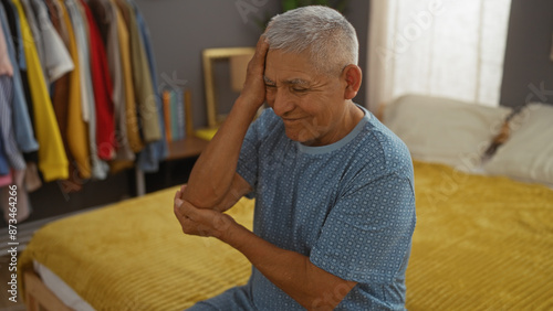 Grey-haired hispanic man sits on a bed in a cozy bedroom, gently massaging his elbow, showcasing a serene moment in a home interior.