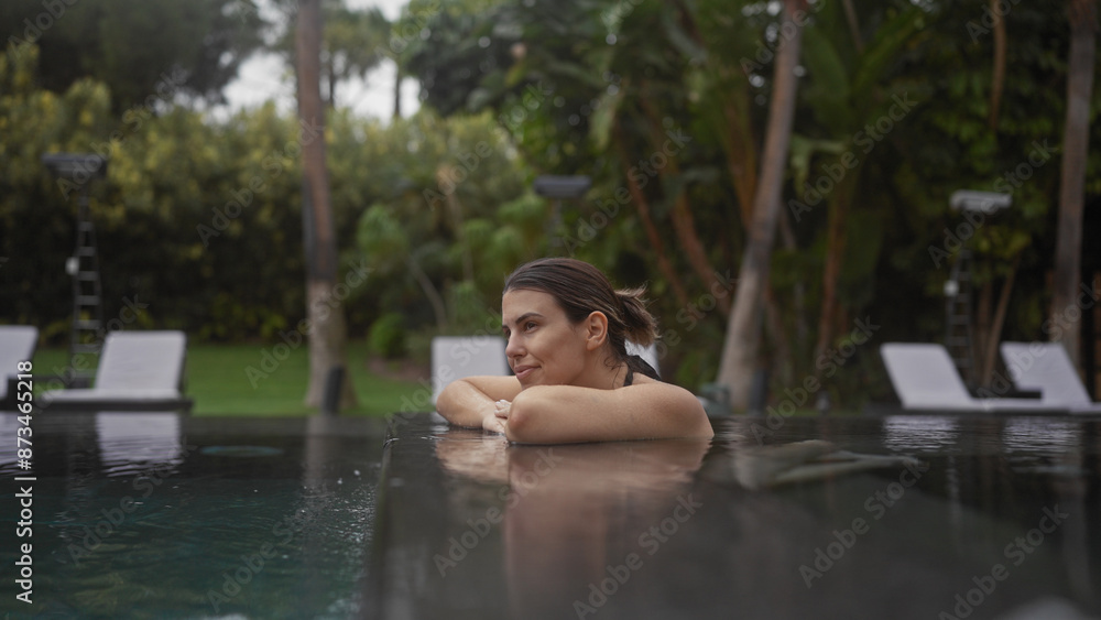 Fototapeta premium Young hispanic woman enjoying a serene moment in an outdoor pool at a bali resort, surrounded by lush tropical greenery.