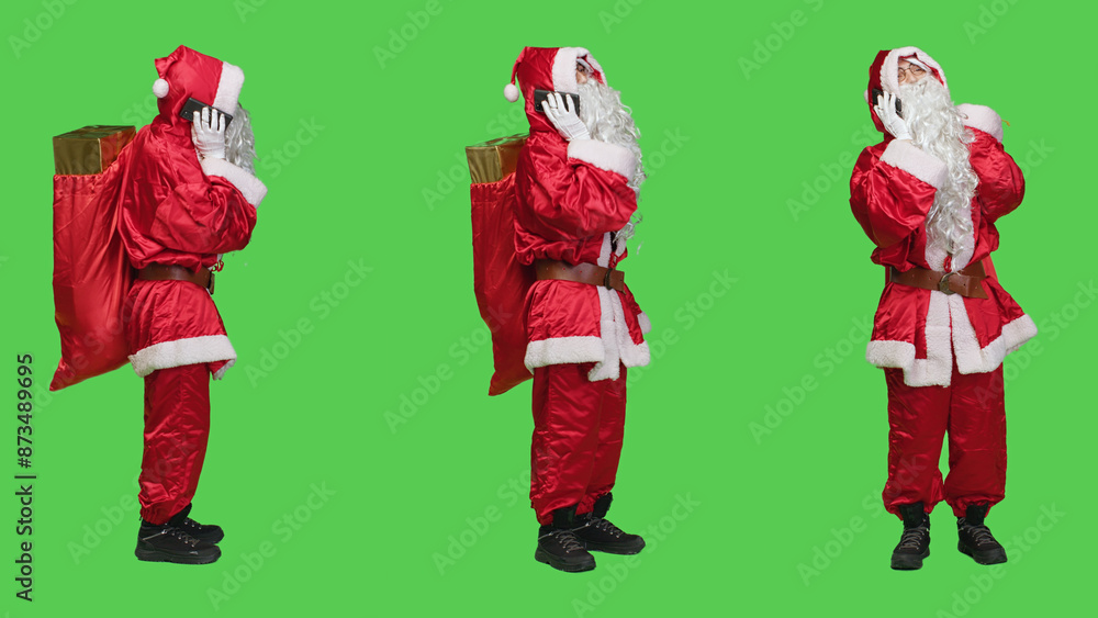 Modern santa claus answers phone call in studio with full body greenscreen, talking on remote chat using smartphone. Father christmas carries sack of presents and toys for holidays. Modern santa claus answers phone call in studio with full body greenscreen, talking on remote chat using smartphone. Father christmas carries sack of presents and toys for holidays.