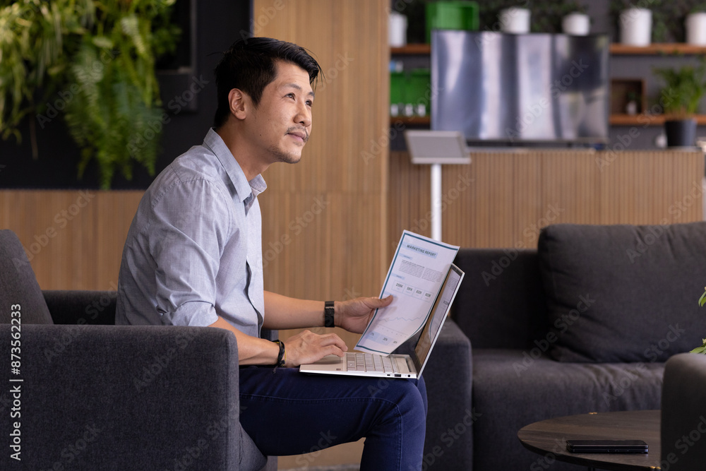 Holding document, man working on laptop in modern office setting Holding document, man working on laptop in modern office setting